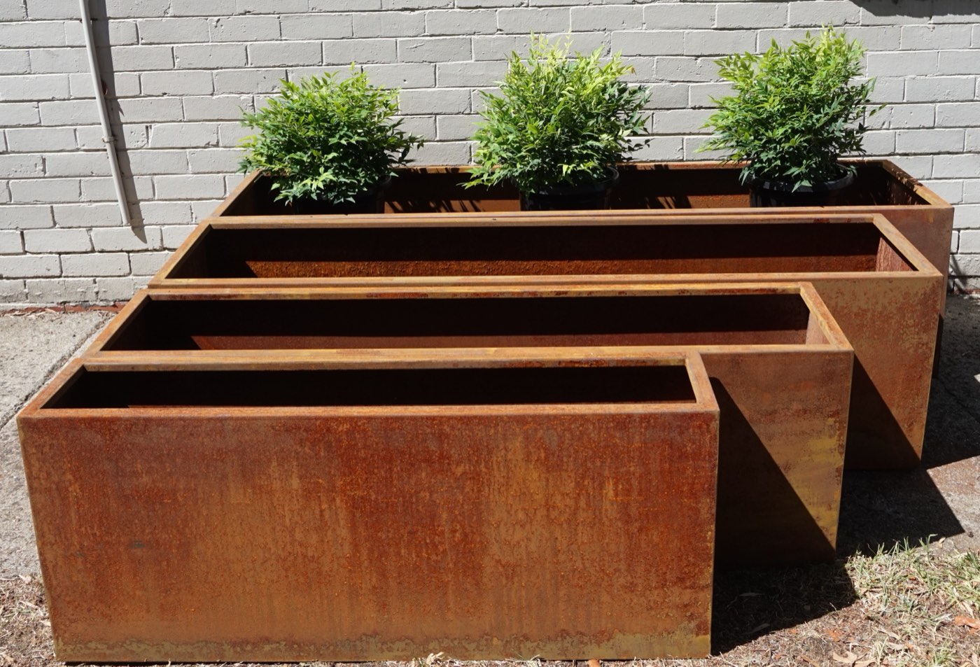 Four corten planters of different sizes nested together outdoors, weathered to an even rust brown, the largest planted with three shrubs.