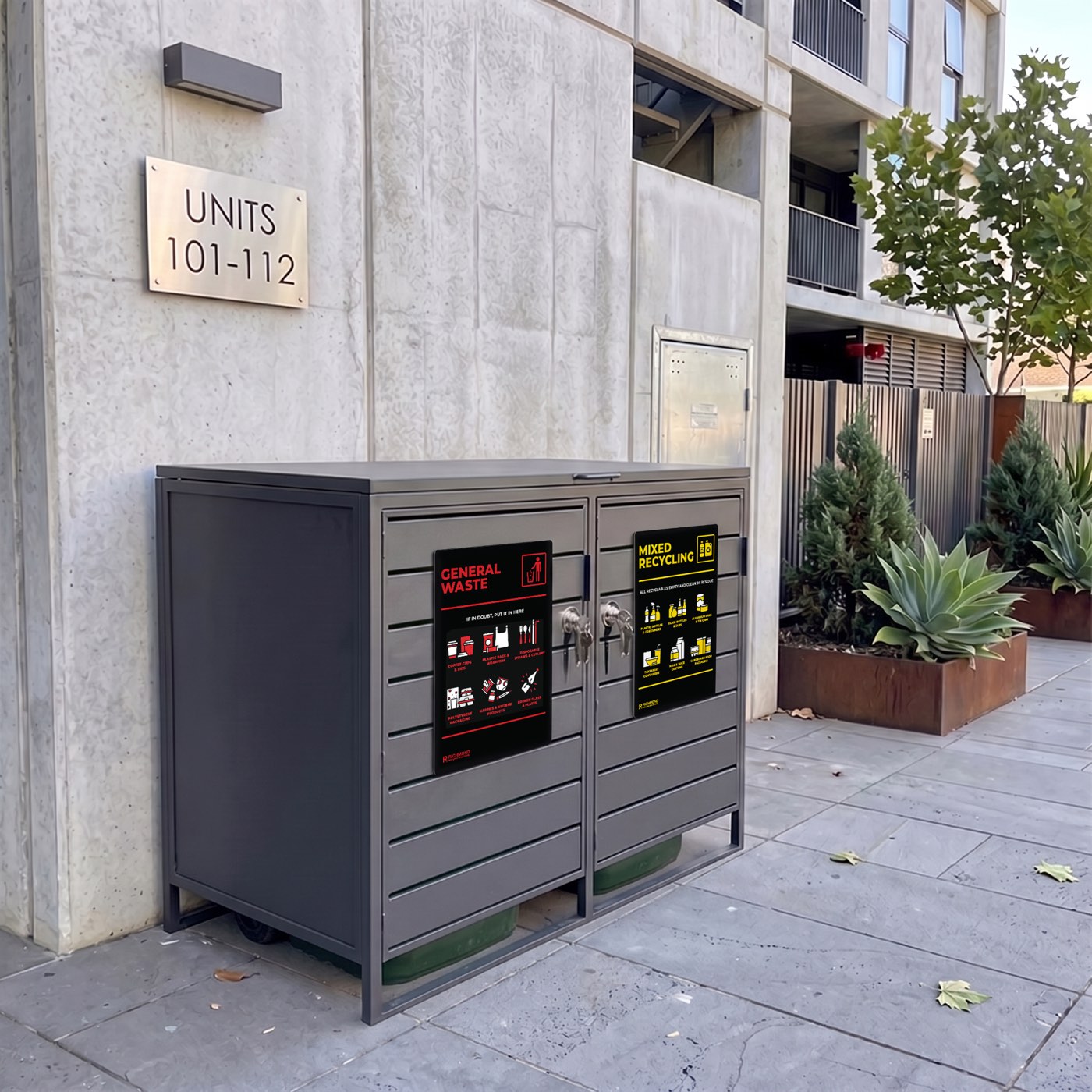 The enclosure installed on a footpath outside an apartment block, with General Waste and Mixed Recycling signage on the doors and green bins visible beneath.